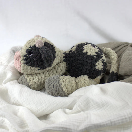 A crocheted plush cow toy resting on soft white and beige fabric bedding. The toy has a beige body with black patches, gray hooves, and a pink nose.