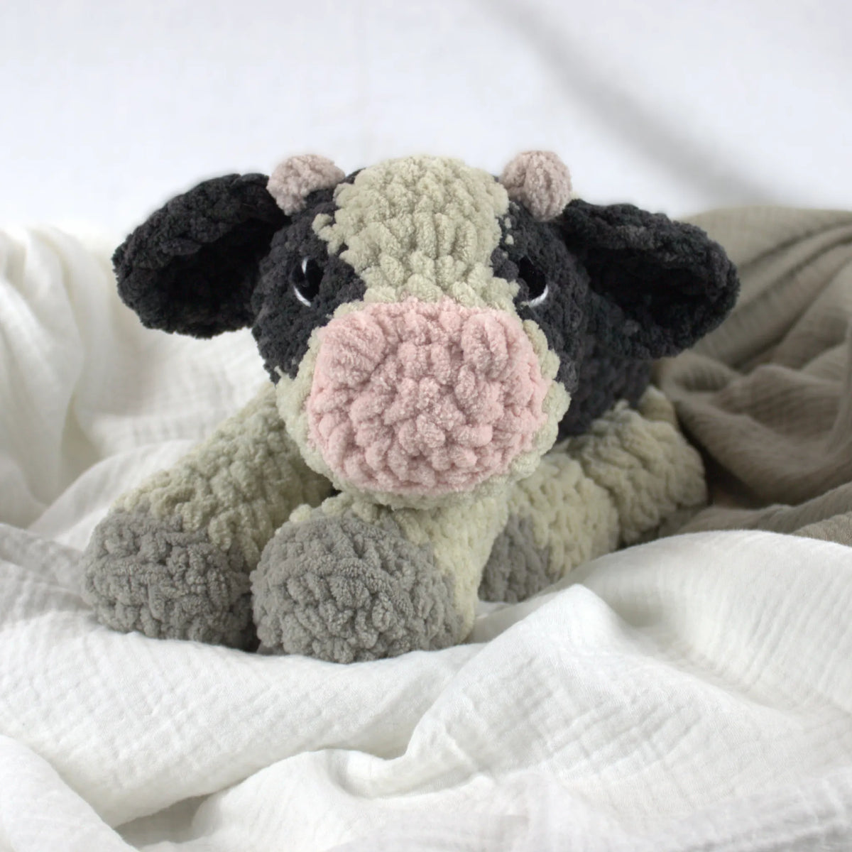 A crocheted plush cow toy with black ears and head, a light beige body, gray hooves, and a pink snout, resting on soft white and beige fabric.