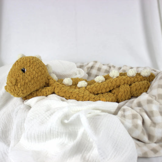 A crocheted plush dinosaur toy in mustard yellow with small cream spikes, resting on white and beige checkered blankets.
