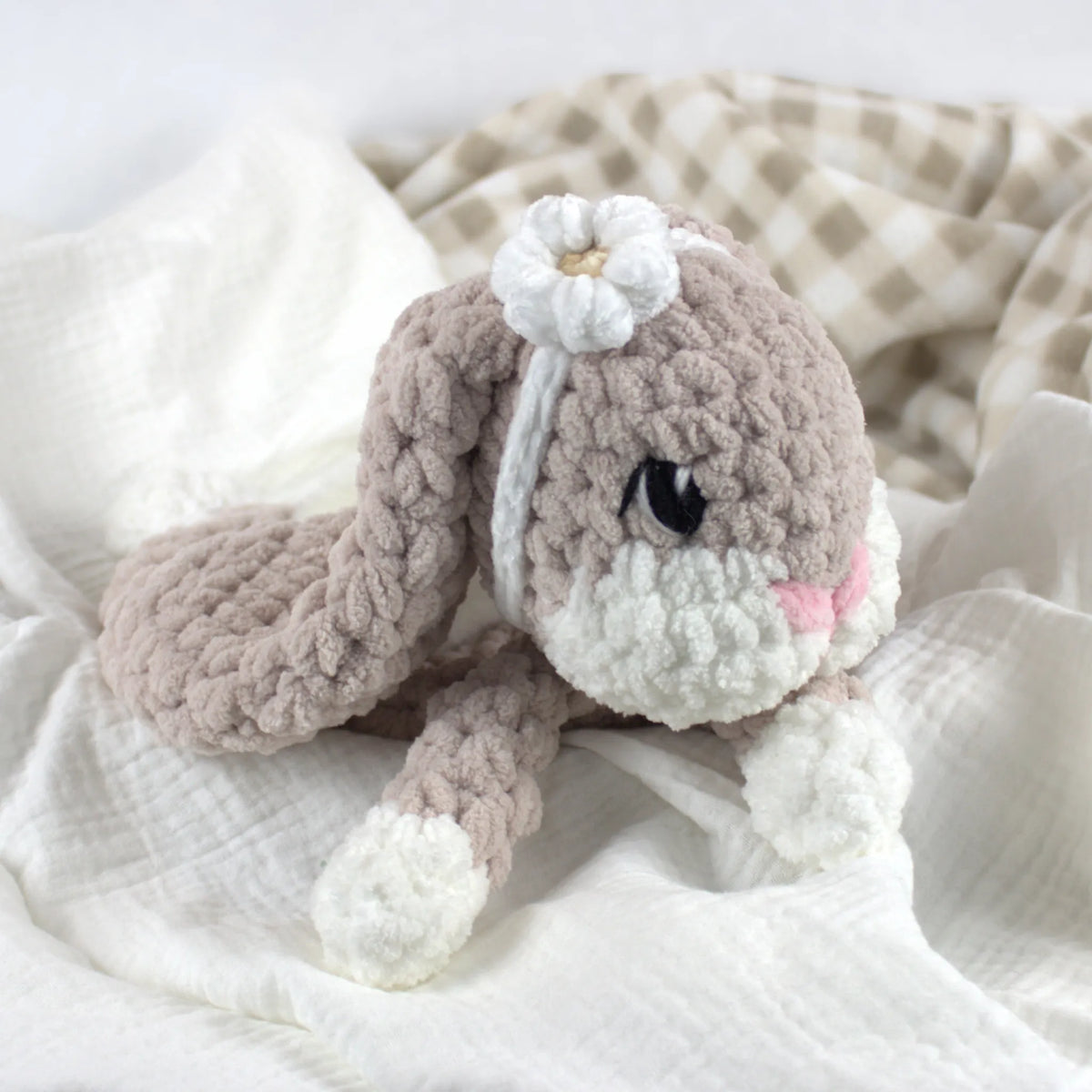 A crocheted plush bunny toy in soft taupe and white, with embroidered black eyes, a pink nose, and a white flower on its head, resting on white fabric with a beige check blanket in the background.