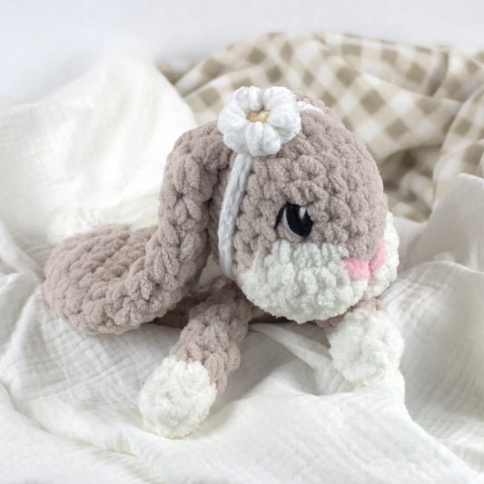 A crocheted plush bunny toy in soft taupe and white, with embroidered black eyes, a pink nose, and a white flower on its head, resting on white fabric with a beige check blanket in the background.