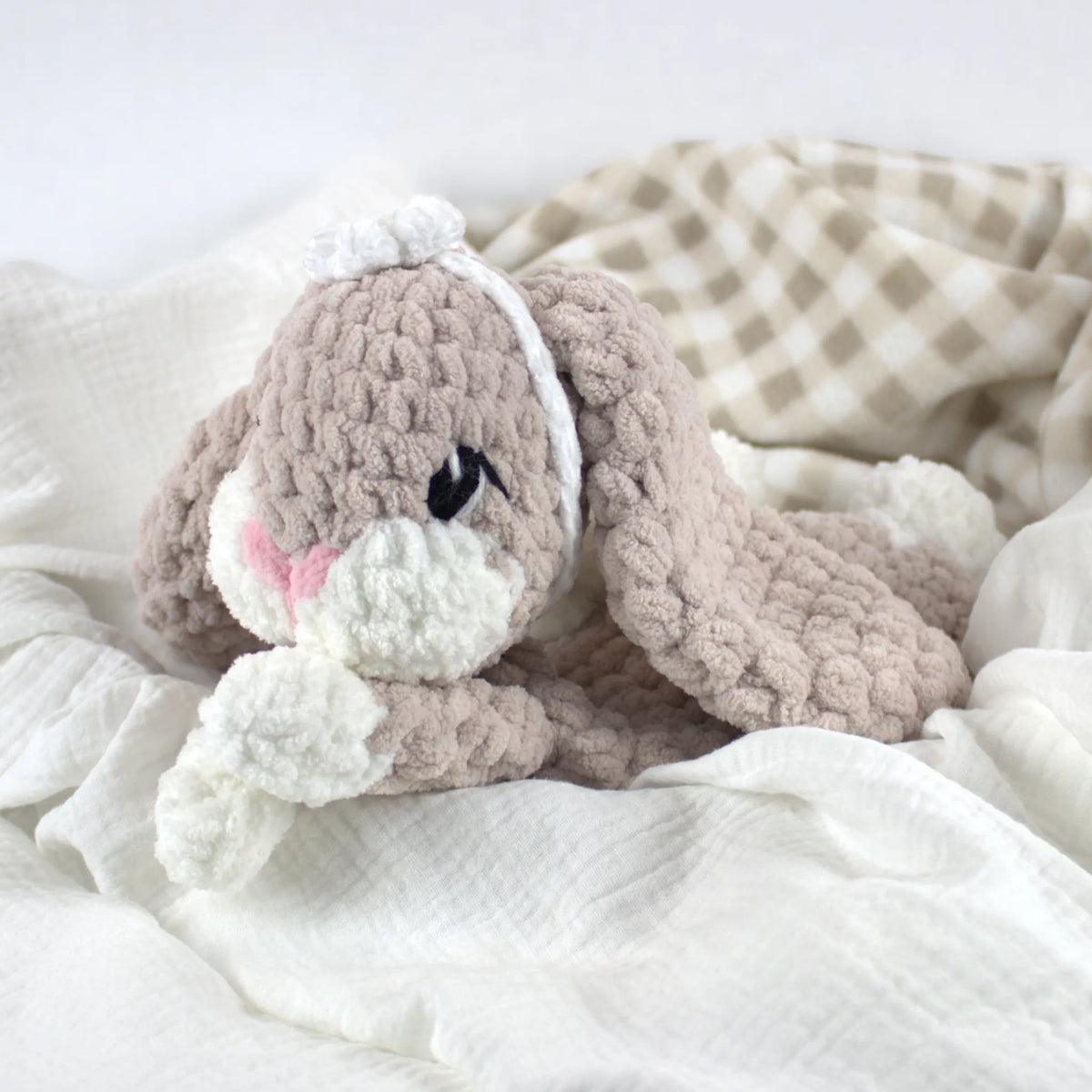 A crocheted bunny plush toy in soft taupe and white, styled on white textured bedding with a beige-and-white check blanket in the background.