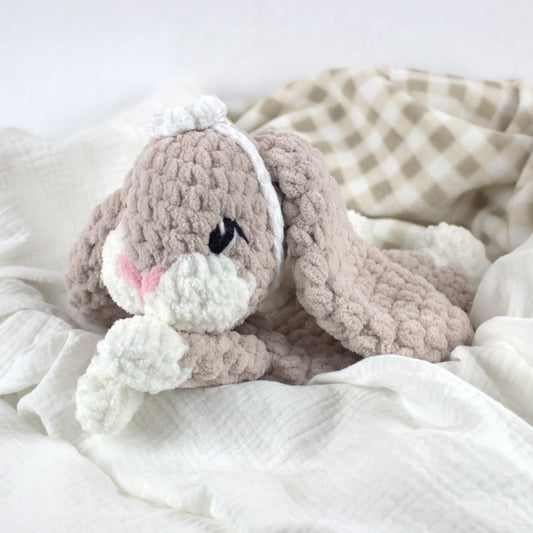 A crocheted bunny plush toy in soft taupe and white, styled on white textured bedding with a beige-and-white check blanket in the background.