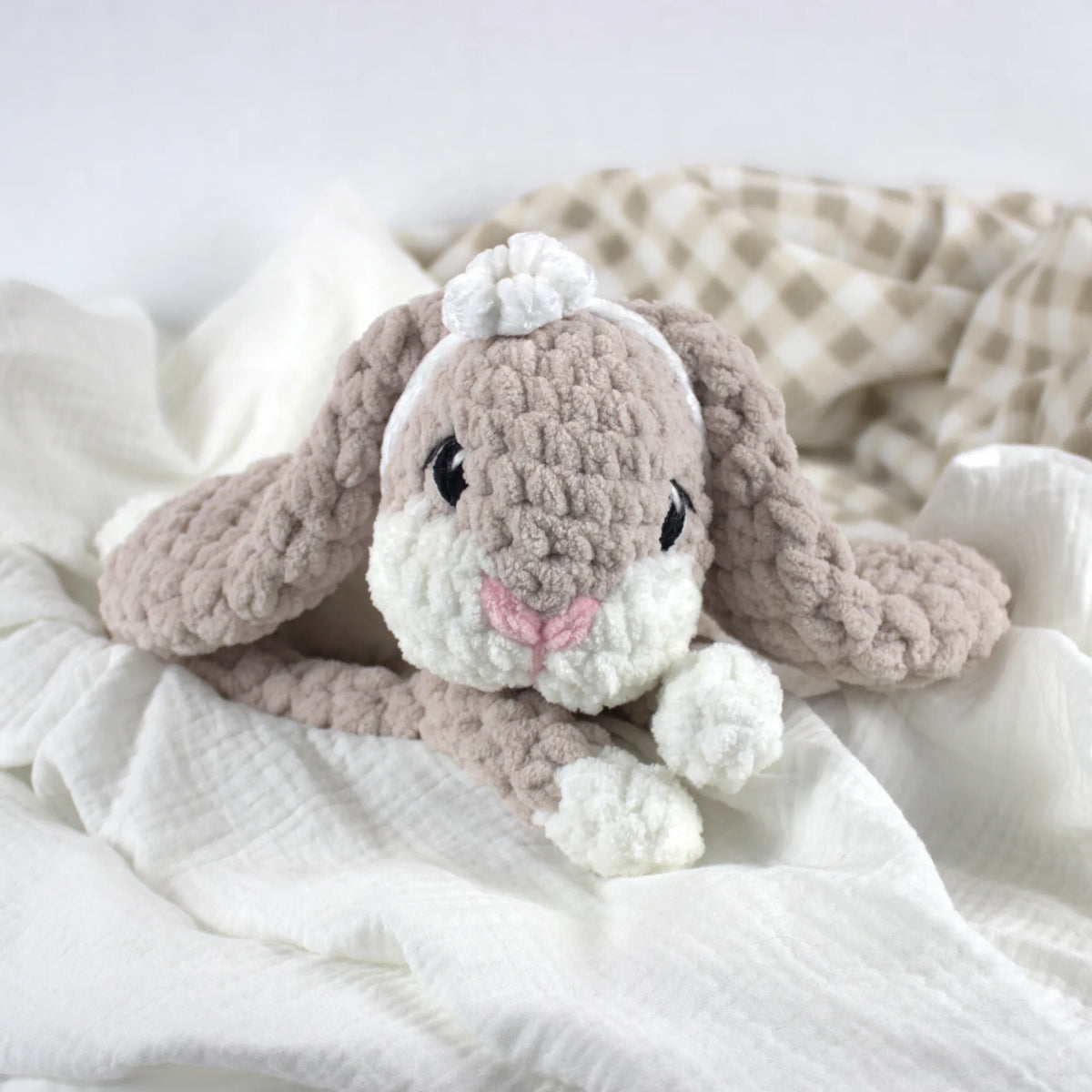 A soft crocheted bunny plush toy in warm taupe and white, styled on white textured fabric with a beige-and-white check blanket in the background.