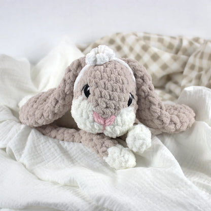 A soft crocheted bunny plush toy in warm taupe and white, styled on white textured fabric with a beige-and-white check blanket in the background.