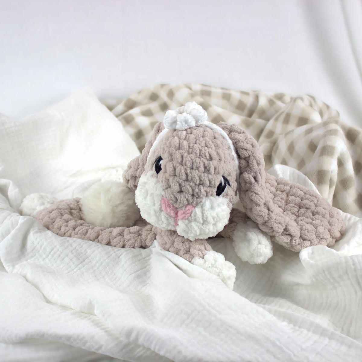 A crocheted bunny plush toy in soft taupe and white, styled on white textured fabric with a beige-and-white striped blanket in the background.