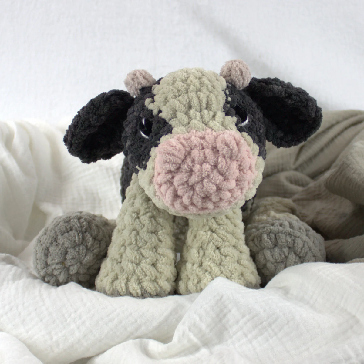 A crocheted plush cow toy with a black and light beige body, pink snout, and gray hooves, resting on white textured fabric.
