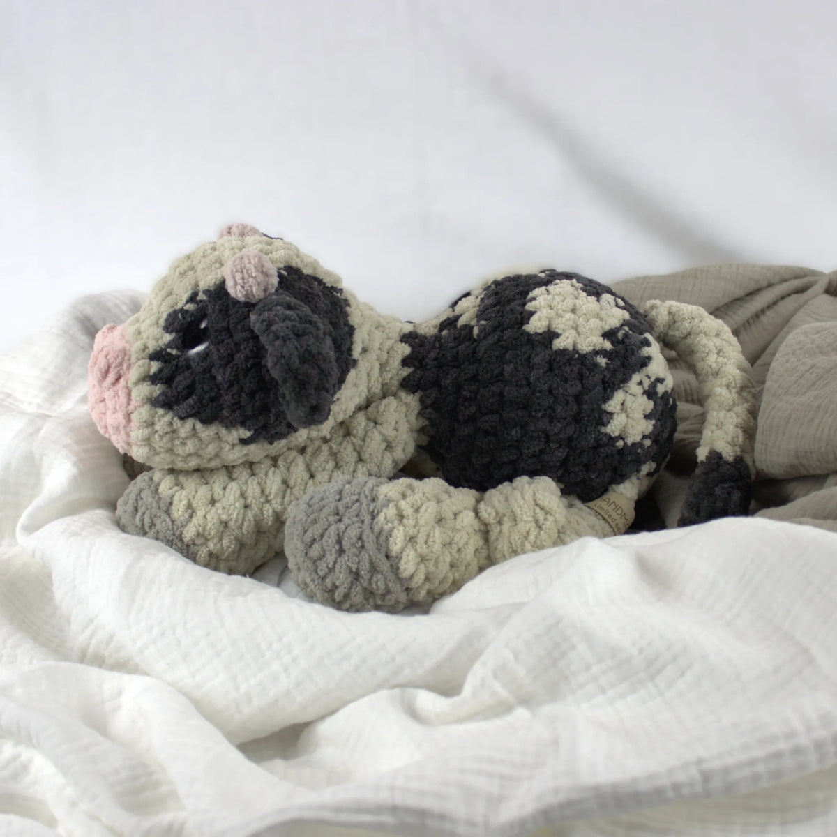 A crocheted plush cow toy resting on soft white and beige fabric bedding. The toy has a beige body with black patches, gray hooves, and a pink nose.