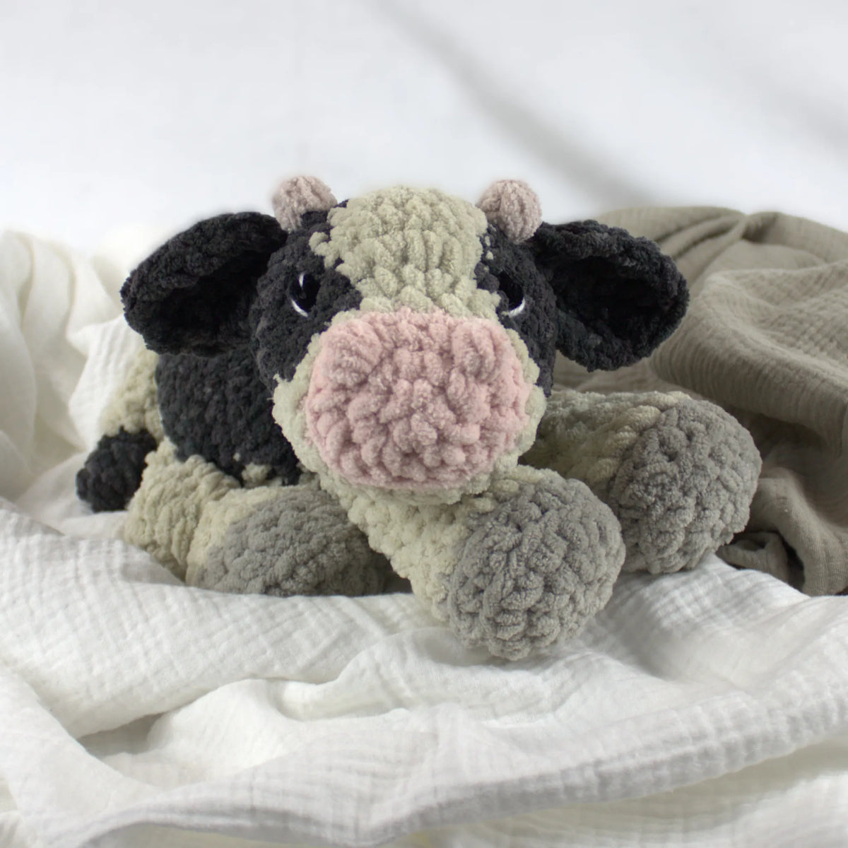 A crocheted plush cow toy with black, beige, and pink colors, resting on soft white and beige fabric.