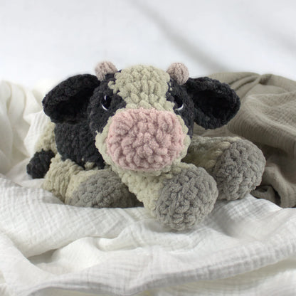 A crocheted plush cow toy with black, beige, and pink colors, resting on soft white and beige fabric.