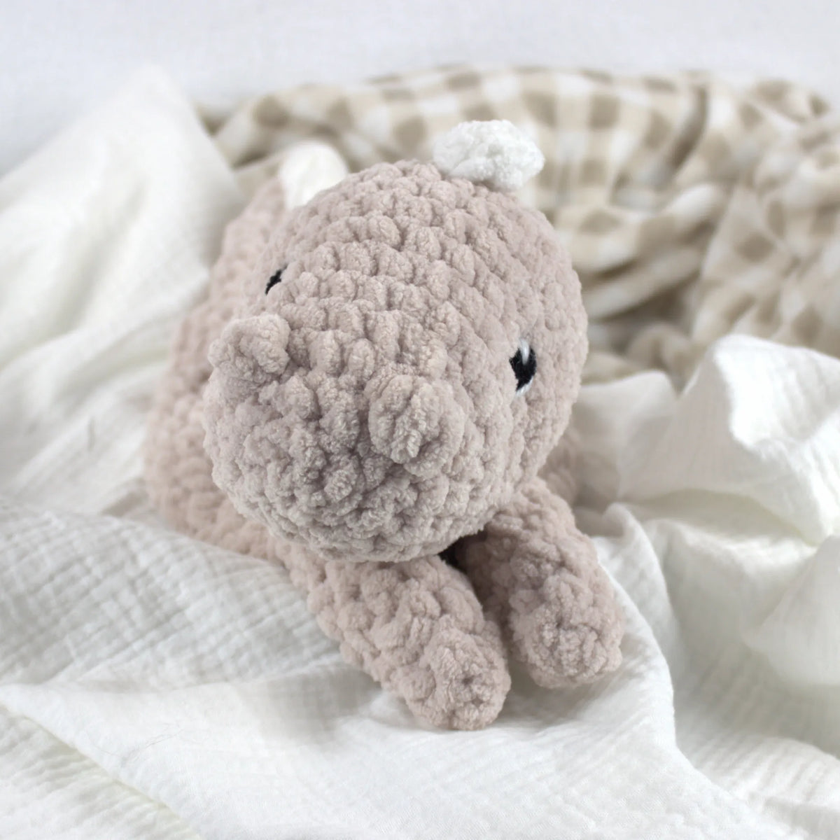 A soft crocheted dinosaur plush toy, resting on white textured fabric with a beige-and-white check blanket in the background.