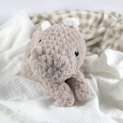 A soft crocheted dinosaur plush toy, resting on white textured fabric with a beige-and-white check blanket in the background.