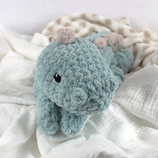 A crocheted plush dinosaur toy in a dusty blue-green color with small beige accents on its ears and a black bead eye, resting on soft white and beige muslin fabric.