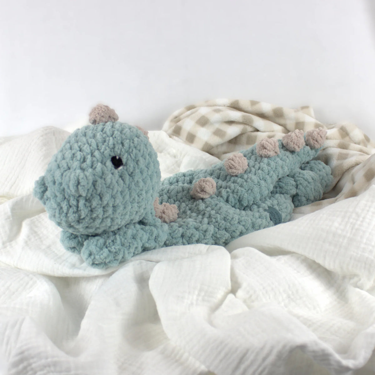 A crocheted dinosaur plush toy in dusty blue with small beige spots, resting on white and beige checkered baby blankets.