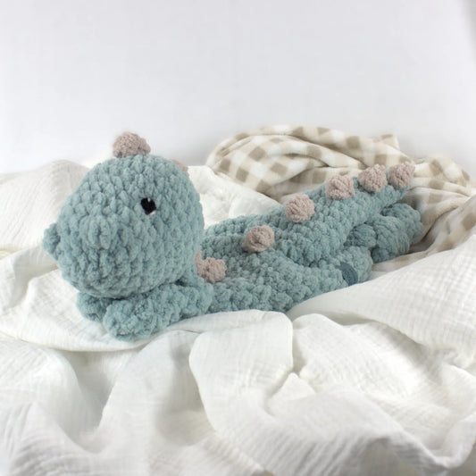 A crocheted dinosaur plush toy in dusty blue with small beige spots, resting on white and beige checkered baby blankets.