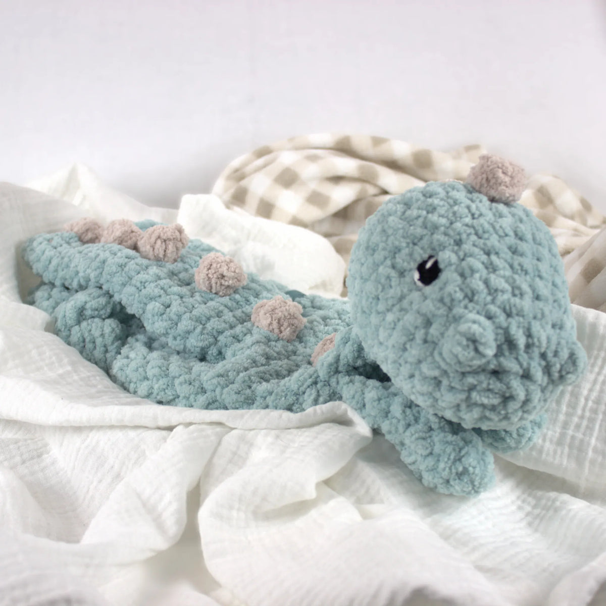 A crocheted dinosaur baby blanket in a dusty teal-blue color with small beige accents on the spikes and head, resting on white gauze fabric.