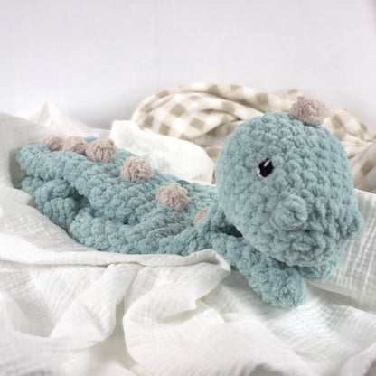 A crocheted dinosaur baby blanket in a dusty teal-blue color with small beige accents on the spikes and head, resting on white gauze fabric.