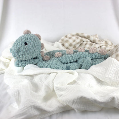 A handmade crocheted dinosaur baby toy in dusty teal blue, styled on soft white muslin gauze fabric with a beige and white checkered blanket.