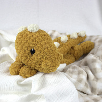 A crocheted plush dinosaur toy in mustard yellow with cream spikes and a black bead eye, resting on white textured fabric.