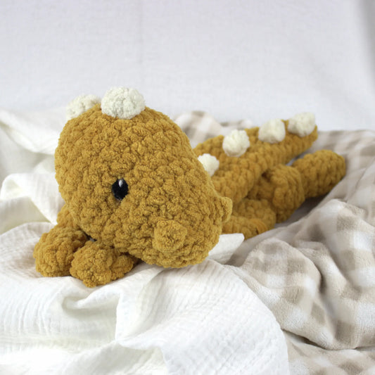 A crocheted plush dinosaur toy in mustard yellow with cream spikes and a black bead eye, resting on white textured fabric.
