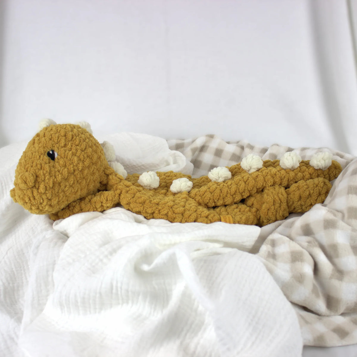 A crocheted plush dinosaur toy in mustard yellow with small cream spikes, resting on white and beige checkered blankets.