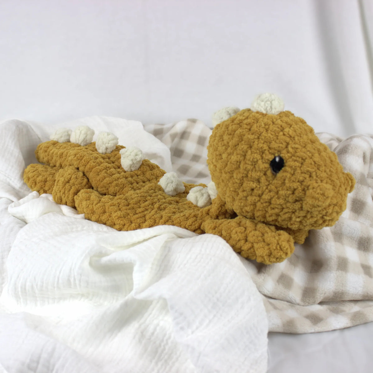 A handmade crocheted plush dinosaur toy in mustard yellow, resting on a white textured muslin cloth and a beige-and-white checkered blanket.