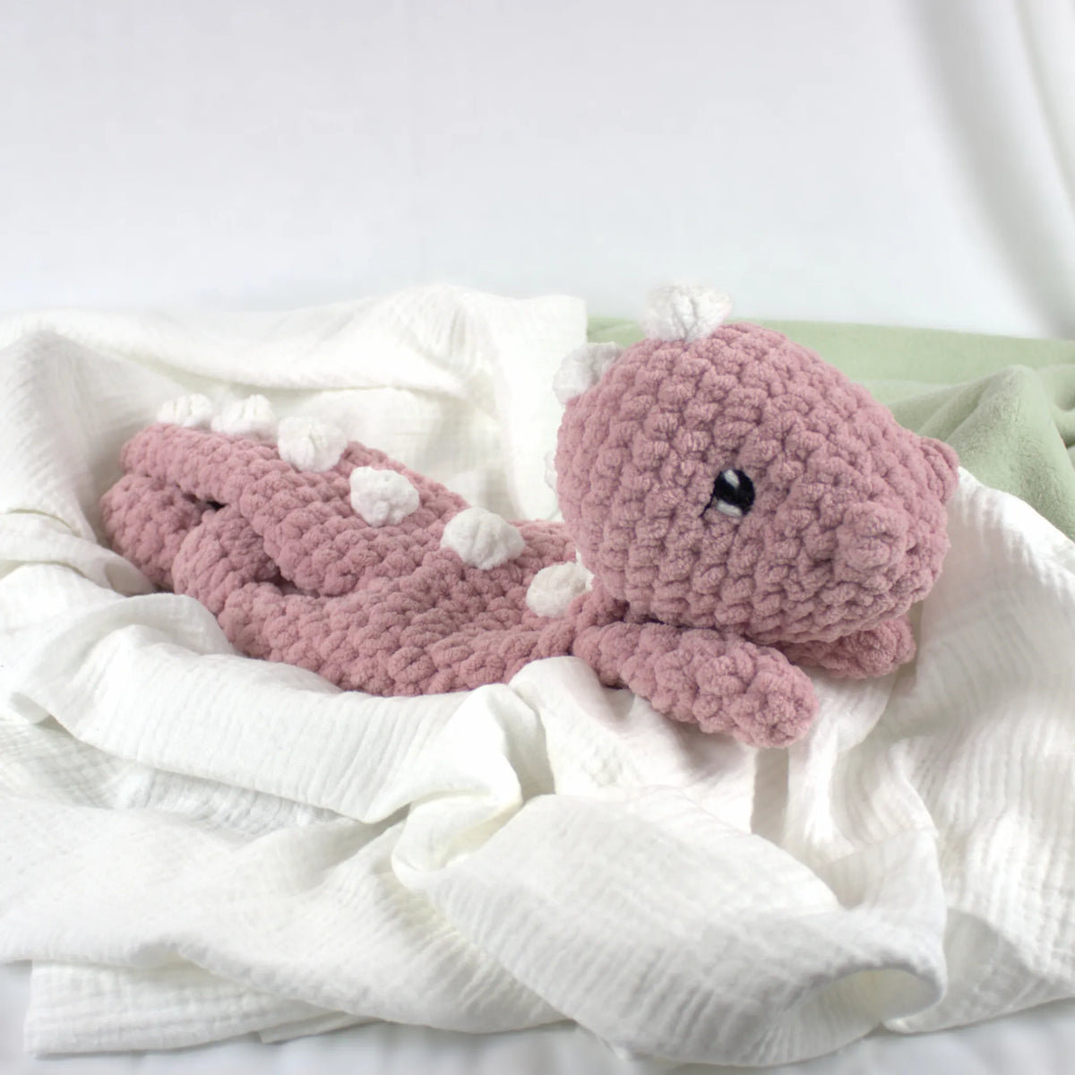 A crocheted pink dinosaur toy with small white accents and a black bead eye, resting on white textured fabric.