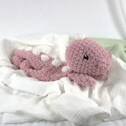 A crocheted pink dinosaur toy with small white accents and a black bead eye, resting on white textured fabric.