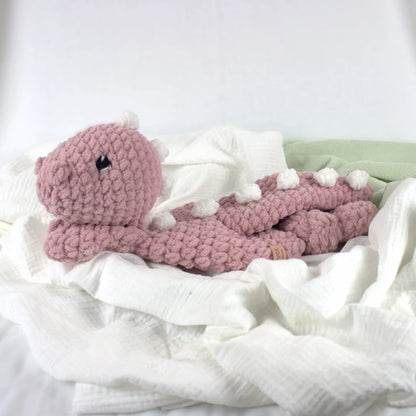 A pink crocheted dinosaur toy resting on soft white muslin fabric with a pale green blanket in the background.