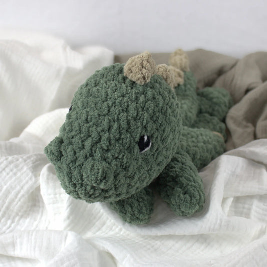 A green crocheted plush dinosaur toy with beige spines, resting on soft white and beige fabric.