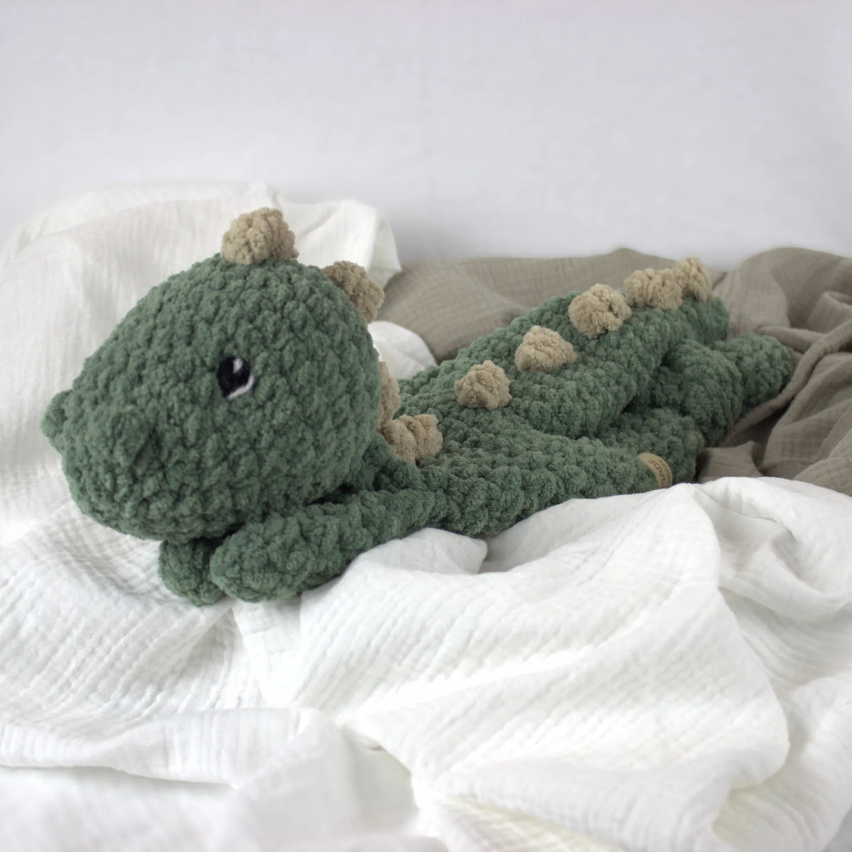 A handmade crocheted plush dinosaur toy in sage green with beige spikes, resting on white and beige textured blankets.