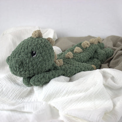 A handmade crocheted plush dinosaur toy in sage green with beige spikes, resting on white and beige textured blankets.