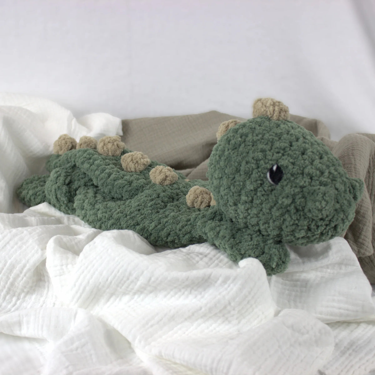A handmade crocheted dinosaur plush toy in sage green with beige spikes, resting on white textured muslin fabric.