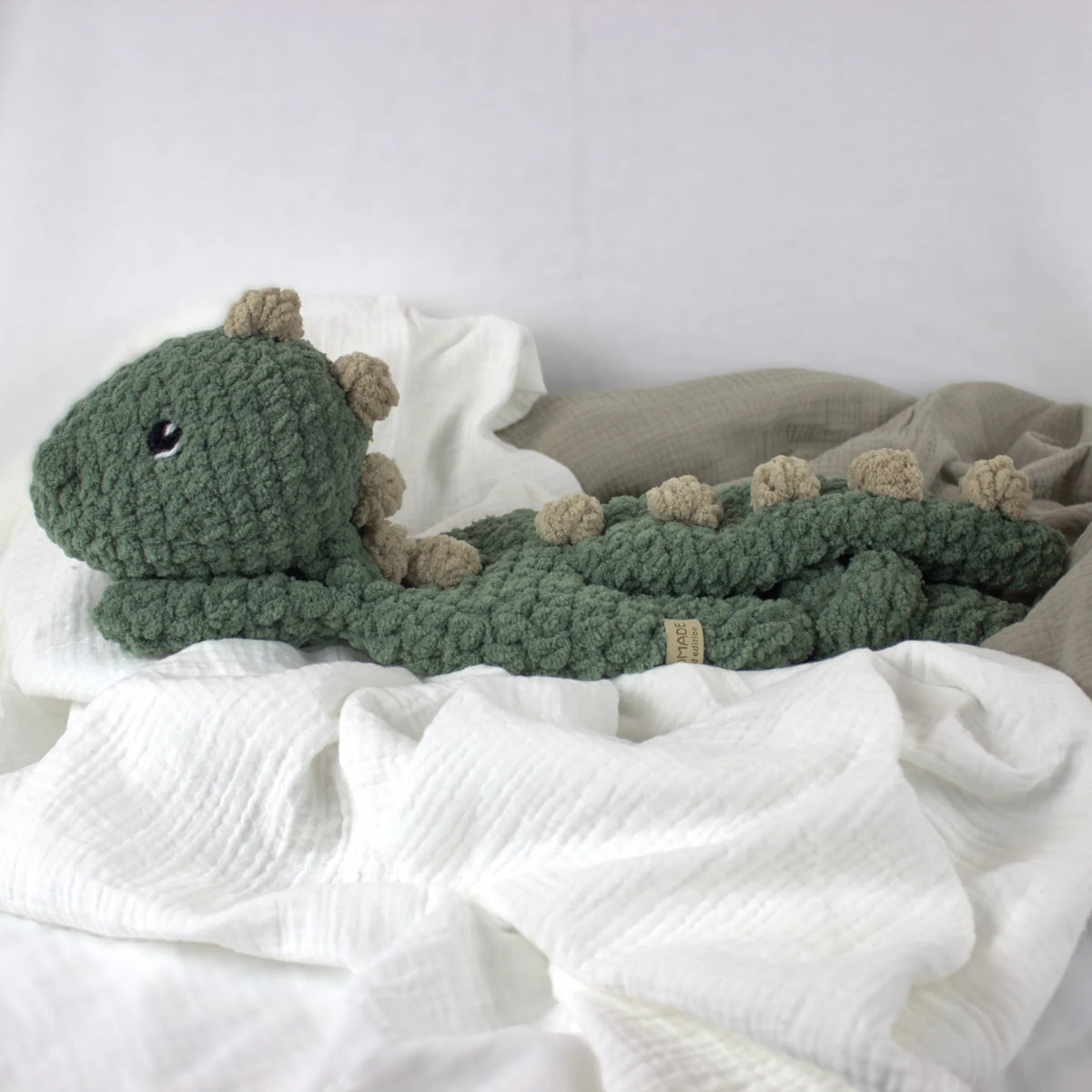 A crocheted plush dinosaur toy in muted green with beige spikes and a small black eye, resting on white and taupe fabric.