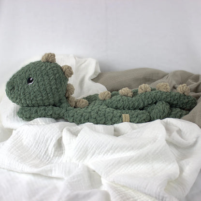 A crocheted plush dinosaur toy in muted green with beige spikes and a small black eye, resting on white and taupe fabric.