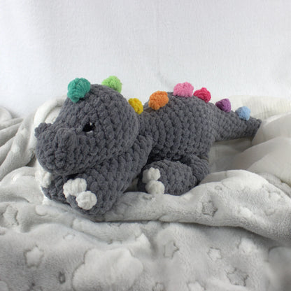 A crocheted plush dinosaur toy in dark gray yarn with colorful spines in green, yellow, orange, pink, purple, and blue, resting on a soft white blanket.