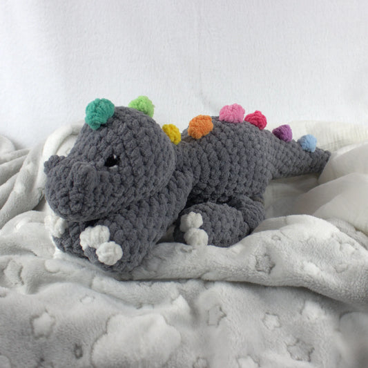 A crocheted plush dinosaur toy in dark gray yarn with colorful spines in green, yellow, orange, pink, purple, and blue, resting on a soft white blanket.
