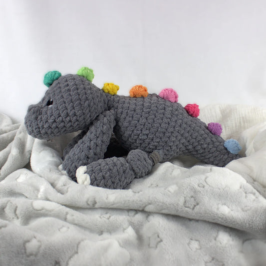 A crocheted dinosaur plush toy in gray with colorful pom-pom spikes, resting on a soft white blanket with star patterns.