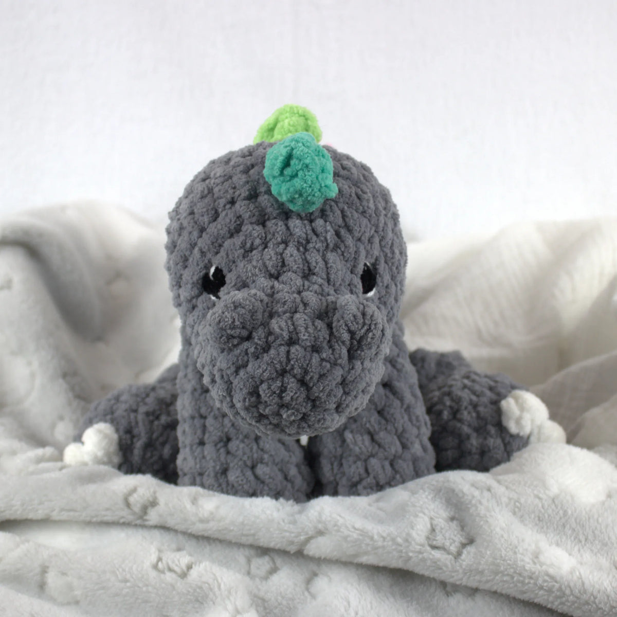 A crocheted plush dinosaur toy in dark gray with small white feet and hands, and a green and light green horn on its head, resting on a soft white blanket.