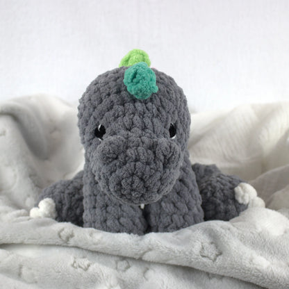 A crocheted plush dinosaur toy in dark gray with small white feet and hands, and a green and light green horn on its head, resting on a soft white blanket.