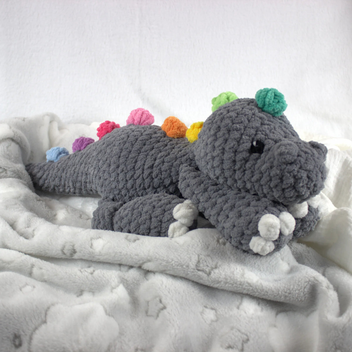 A crocheted plush dinosaur toy in dark gray, resting on a white star-patterned blanket against a white background. The dinosaur features colorful accents on its back and head, including green, pink, orange, yellow, blue, and purple.