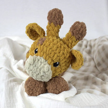 A crocheted plush giraffe toy with a mustard yellow body, brown ossicones and legs, and a cream-colored snout, resting on white and beige textured blankets.