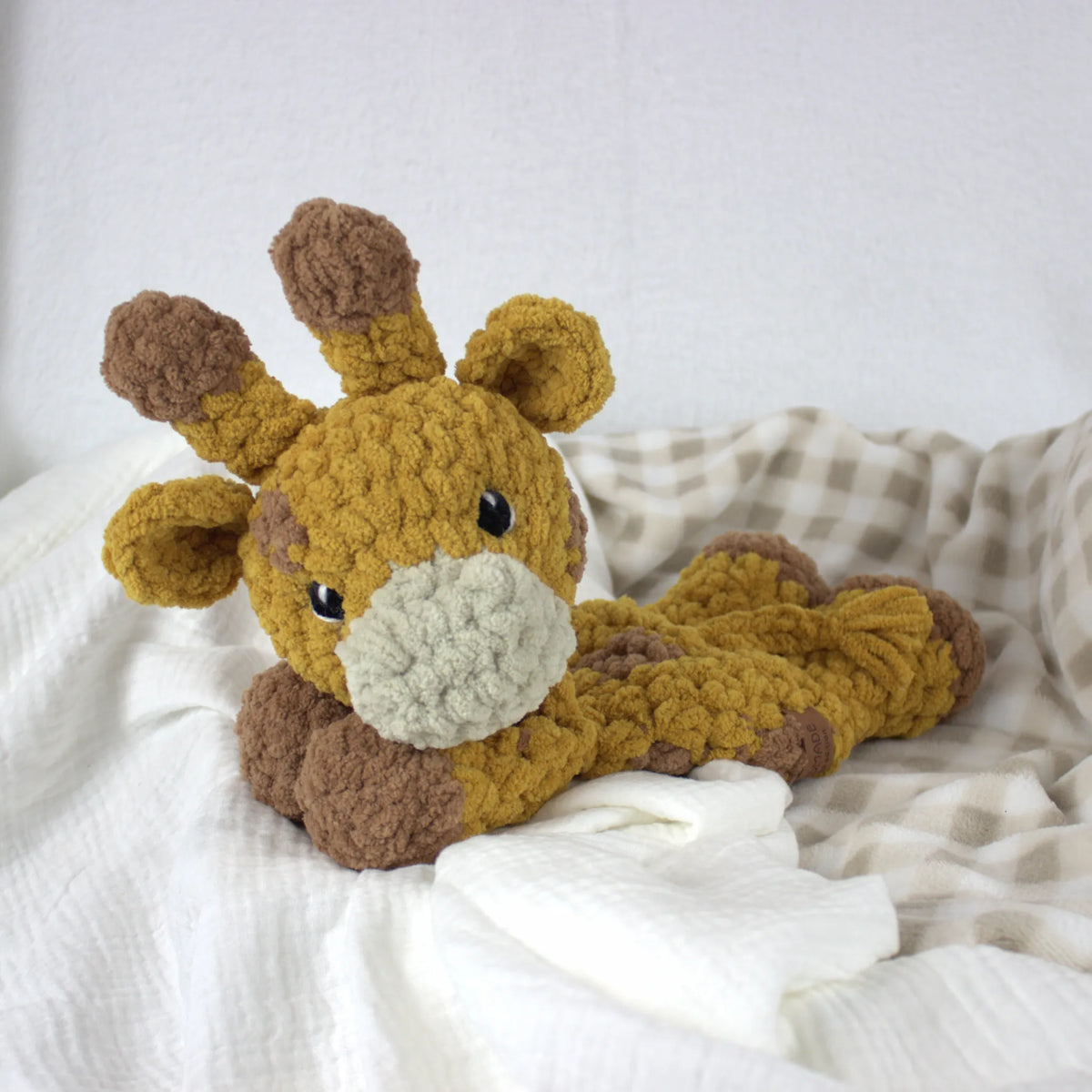 A crocheted plush giraffe toy in mustard yellow and brown, lying on white and beige checkered bedding.