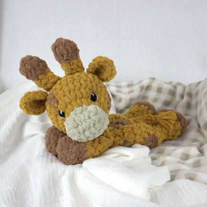 A crocheted plush giraffe toy in mustard yellow and brown, lying on white and beige checkered bedding.