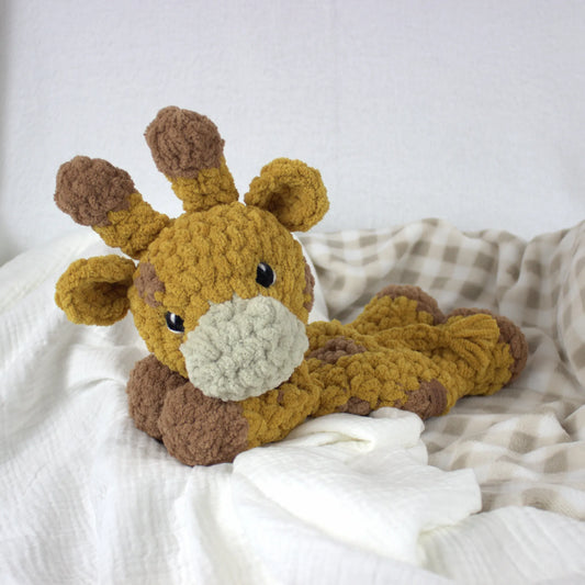A crocheted plush giraffe toy in mustard yellow and brown, lying on white and beige checkered bedding.