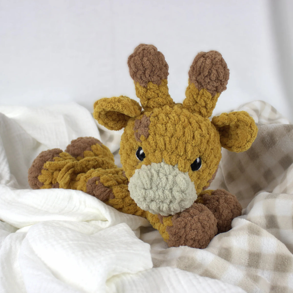 A crocheted plush giraffe toy in mustard yellow and brown yarn, resting on soft white and beige plaid blankets.