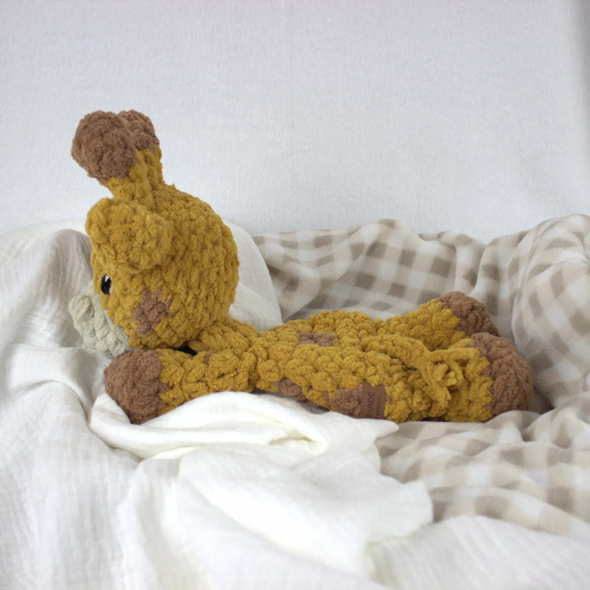 A handmade crocheted giraffe plush toy in mustard yellow and brown, resting on white and beige checkered blankets.