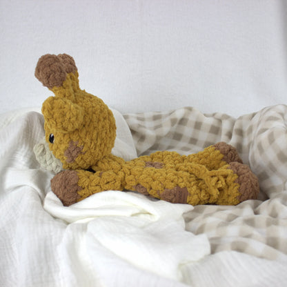 A handmade crocheted giraffe plush toy in mustard yellow and brown, resting on white and beige checkered blankets.