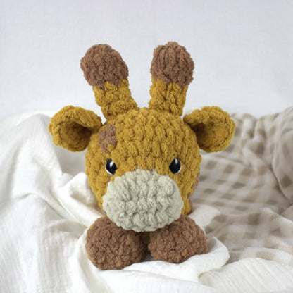 A handmade crocheted giraffe toy with a mustard yellow body, brown ossicones and legs, and a light gray snout, resting on white and beige textured blankets.
