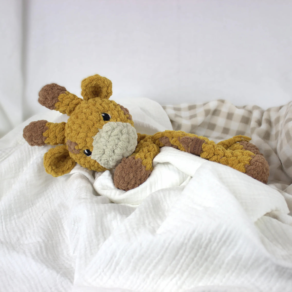 A crocheted plush giraffe toy resting on white textured bedding with a beige-and-white check blanket in the background.